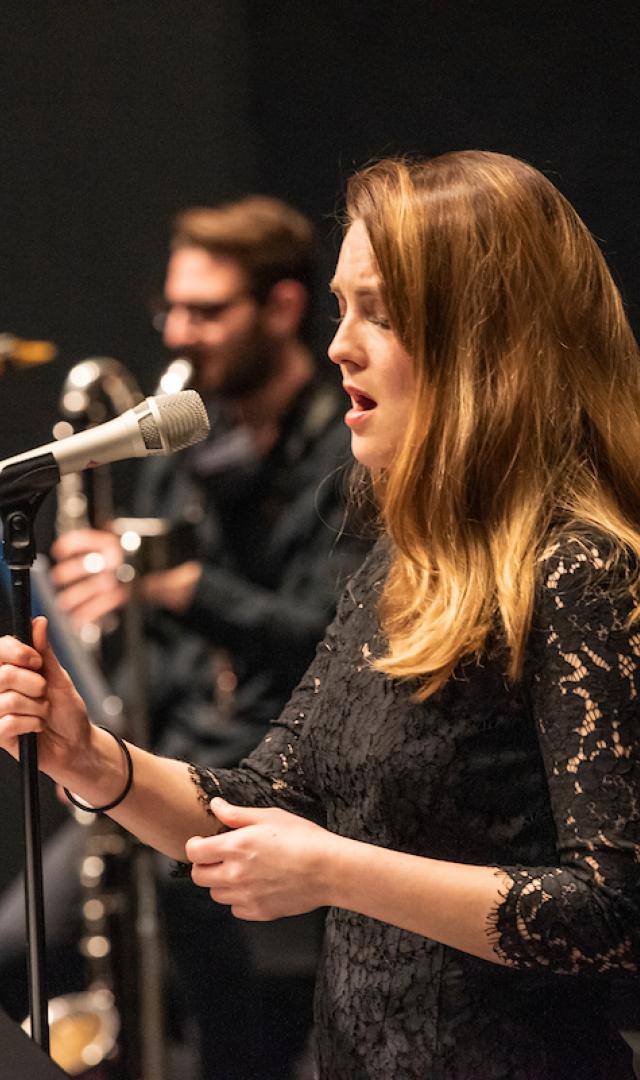 A jazz singer closes her eyes and sings into a microphone, a saxophone player in the background