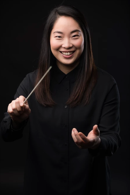 Holly Hyun Choe smiles and holds her conducting baton. She is wearing black and there is a black background.