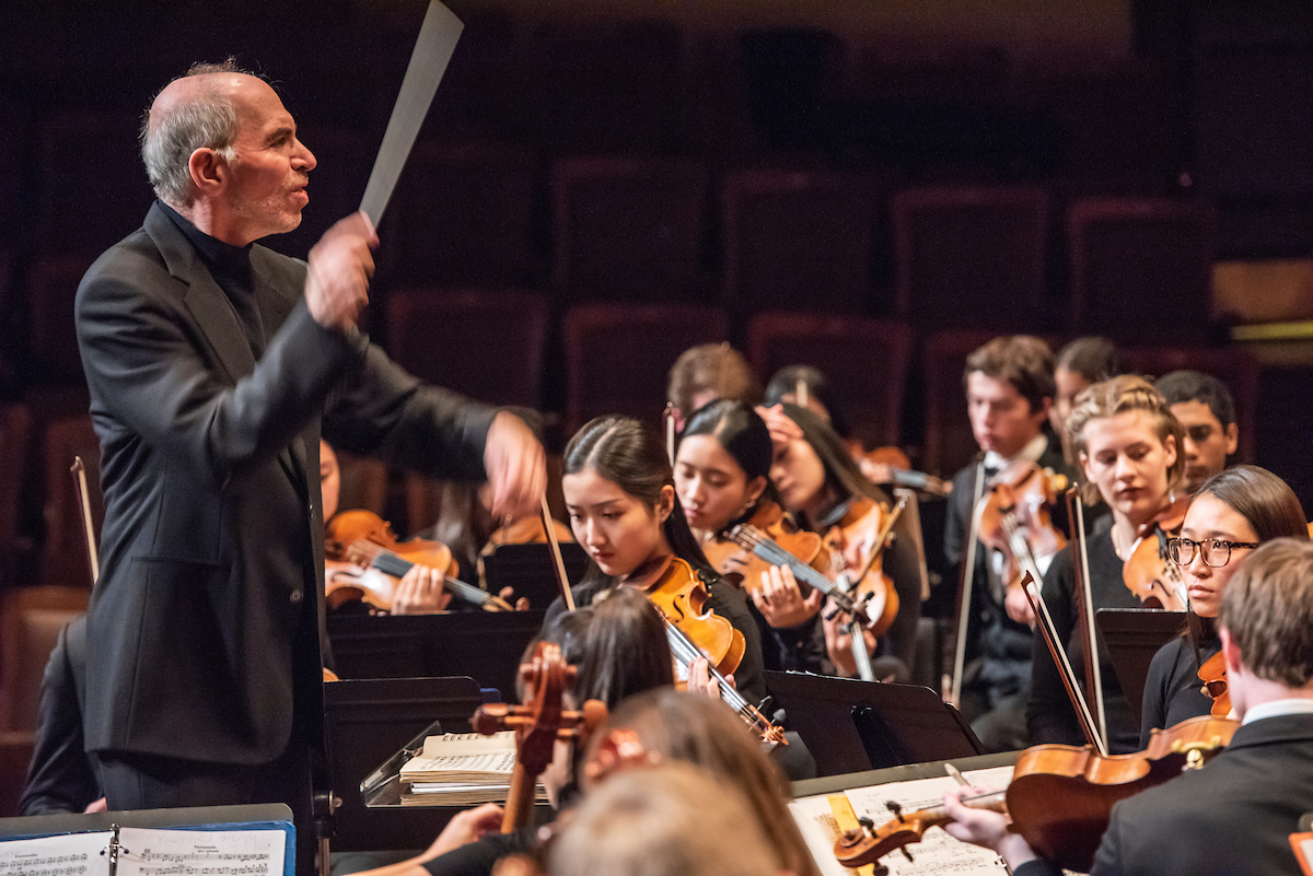 Action shot of David Loebel with conductor baton in motion, while members of the Youth Philharmonic Orchestra play.