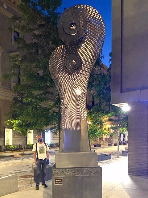 A large statue, approximately 20 feet high, with two scroll shapes like those at the end of a string instrument. The statue is metal, three-dimensional, and lit up from within. It stands at the corner of Gainsborough and St. Botolph streets. In this photo, a man stands at the foot of the statue looking up at it. He is wearing a face mask.