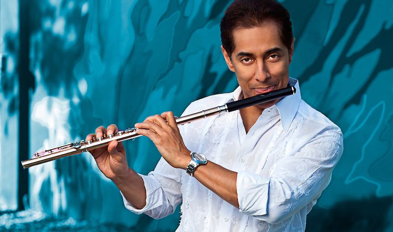 Nestor Torres smiles at the camera while posing with his flute in front of a blue background