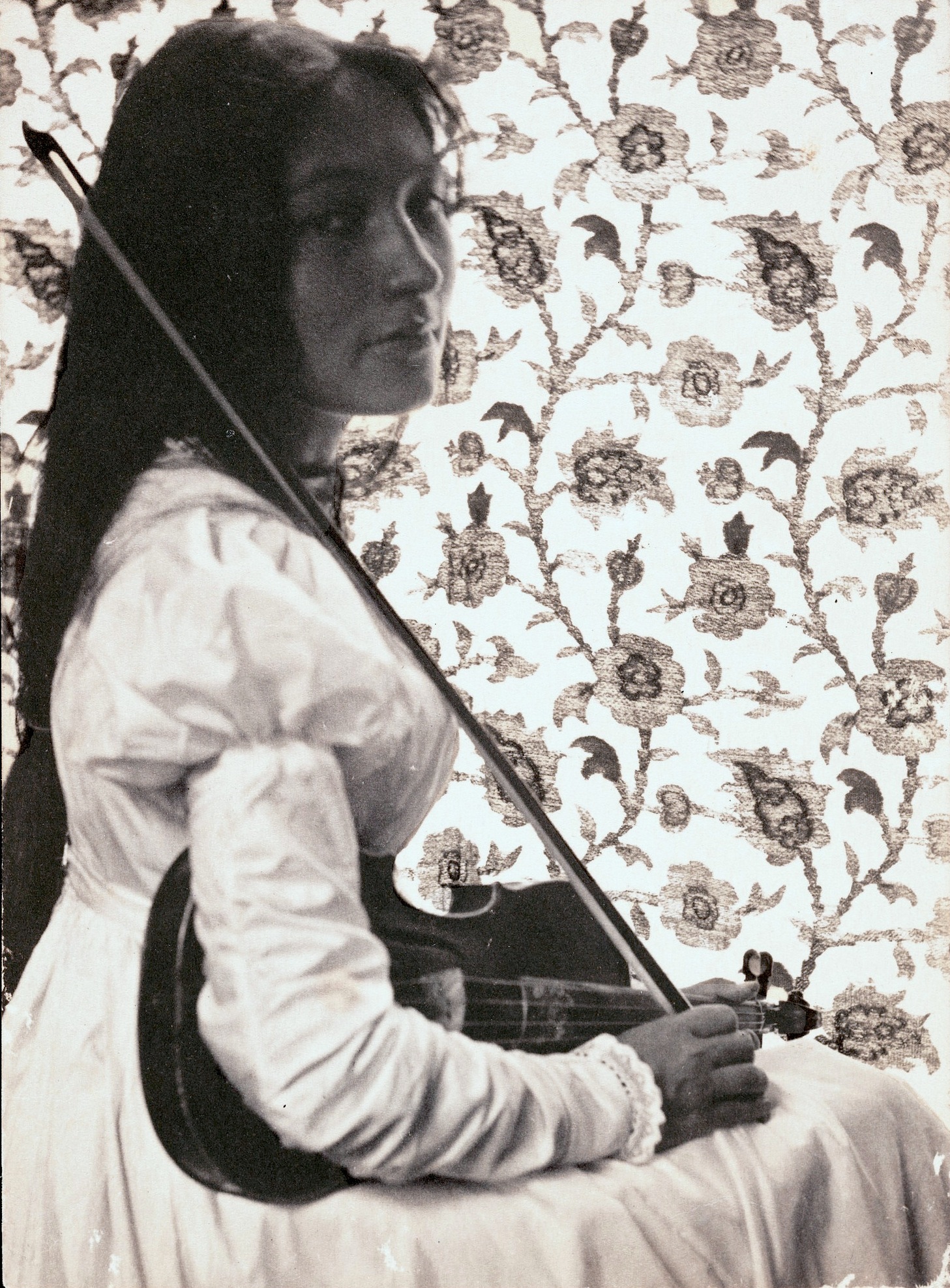 Portrait of Zitkala-Sa, wearing a white puff-sleeve dress, looking over her shoulder and holding her violin. The background is floral wallpaper.