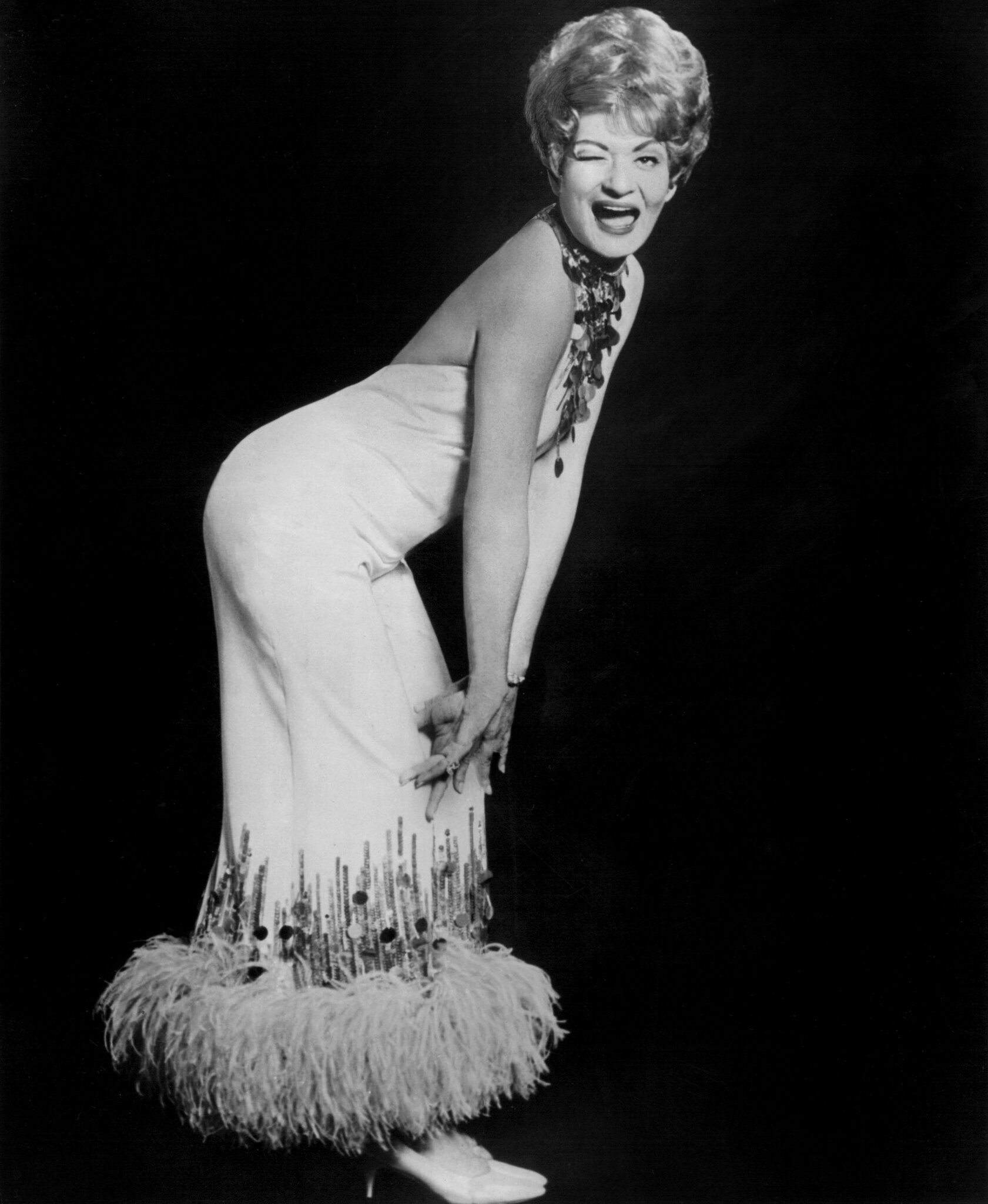 Black and white photo of Rusty Warren in the 1960s. She is bending forward with her hands on her knees, wearing a glamorous dress and winking.
