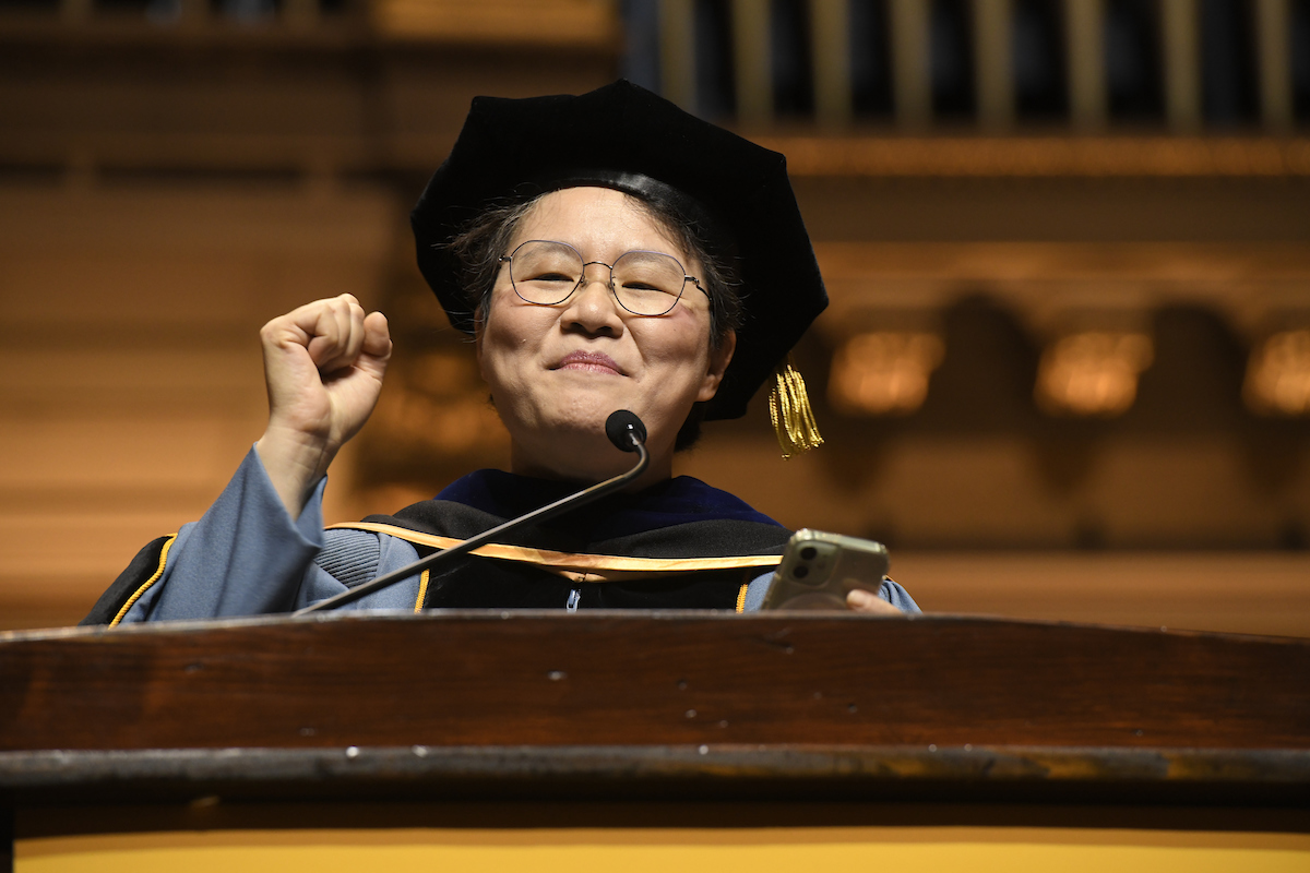 Mei-Ann Chen wearing doctoral regalia, at the podium, clasps her fist and smiles