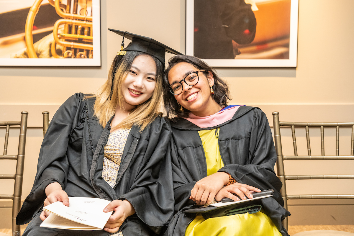 Two graduates lean their heads together and smile while wearing caps and gowns