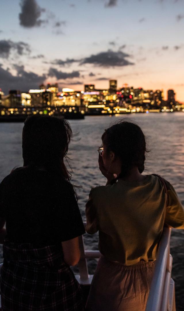 Two students look out at the Boston Harbor and city skyline as the sun sets