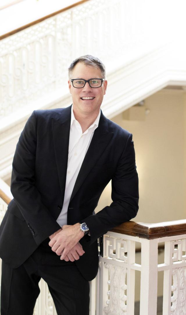 Chad Smith wears a suit and glasses and smiles at the viewer, with a white decorative staircase behind him