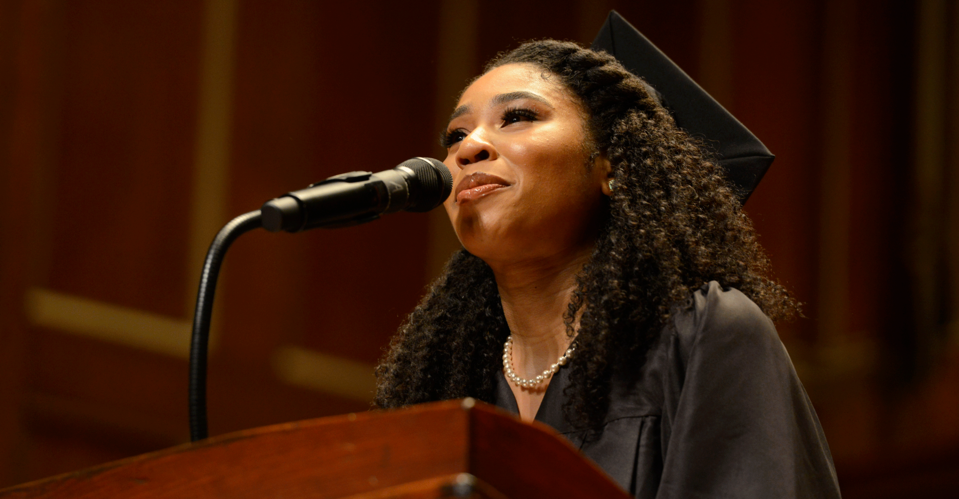 Darynn Dean stands at a podium wearing a graduation cap and gown. She smiles and looks into the distance while speaking into a microphone.