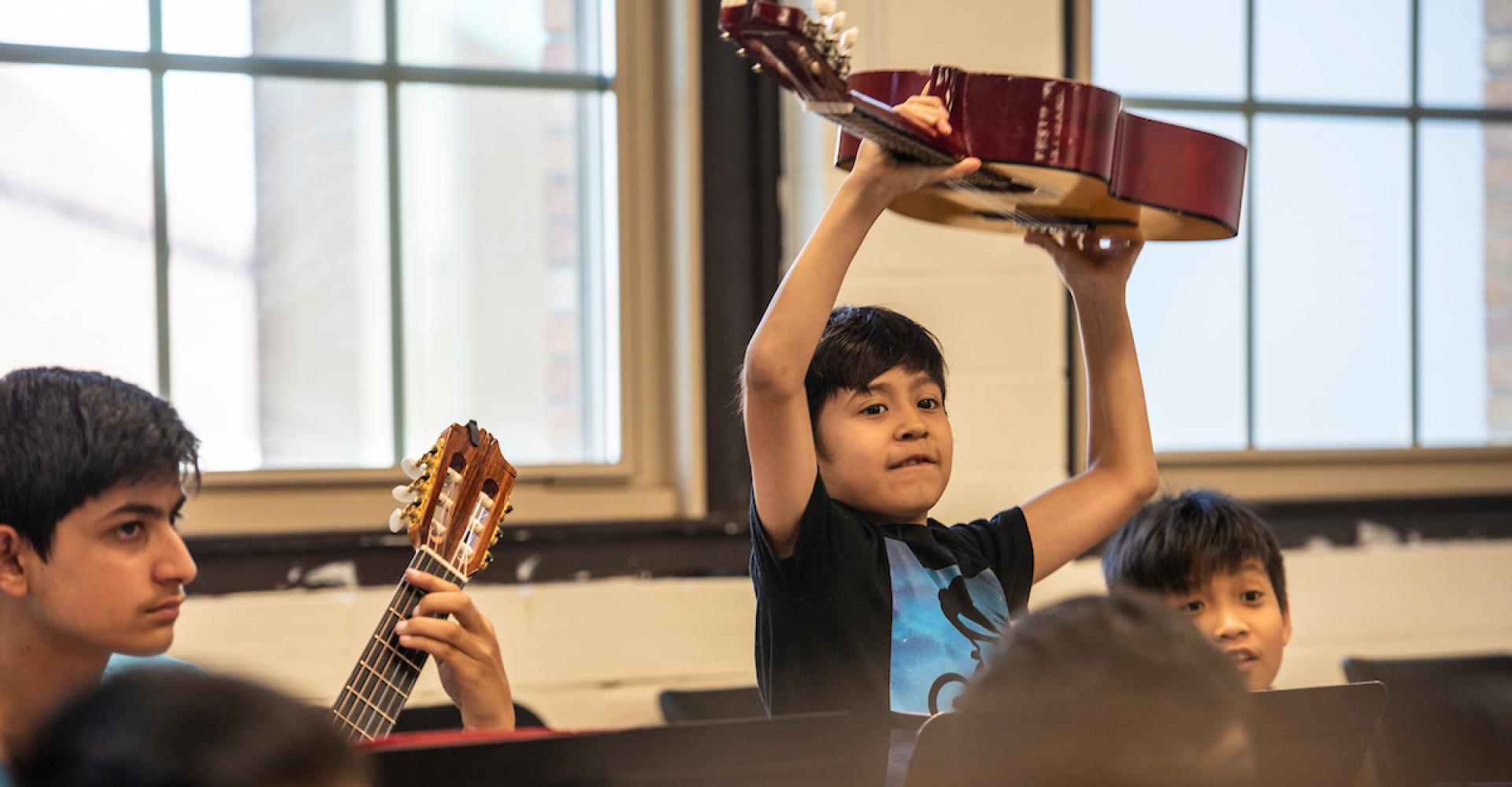 A young guitarist holds his guitar over his head in a group class.