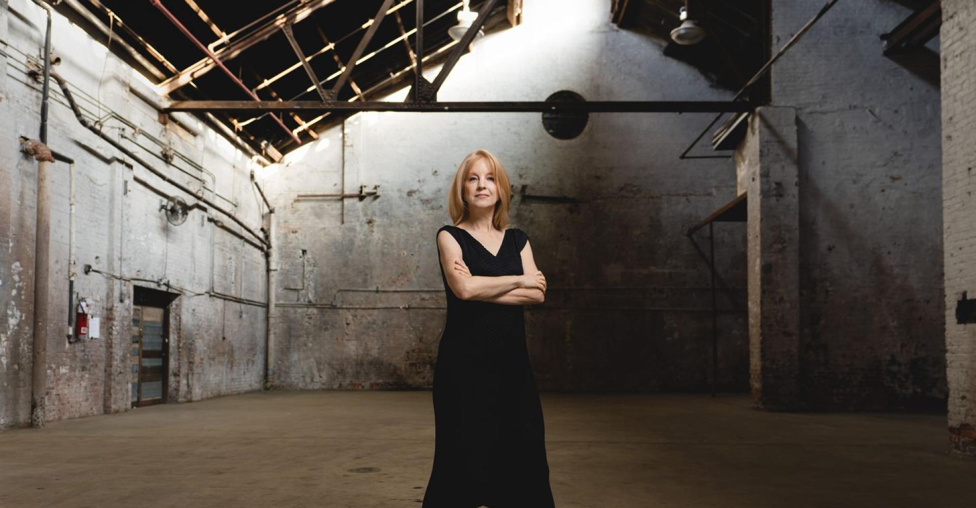 Maria Schneider stands in a warehouse or barn, looking at the camera with arms crossed.