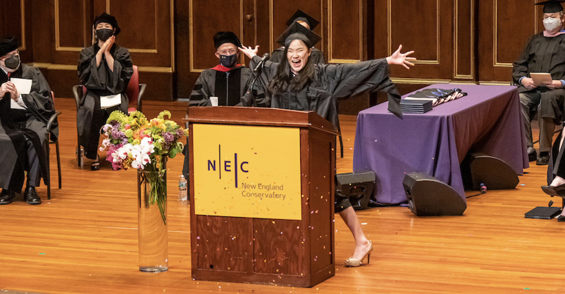 Esther Tien, wearing a graduation cap and gown, opens her arms wide and flings confetti while speaking at the podium at Commencement 2021.