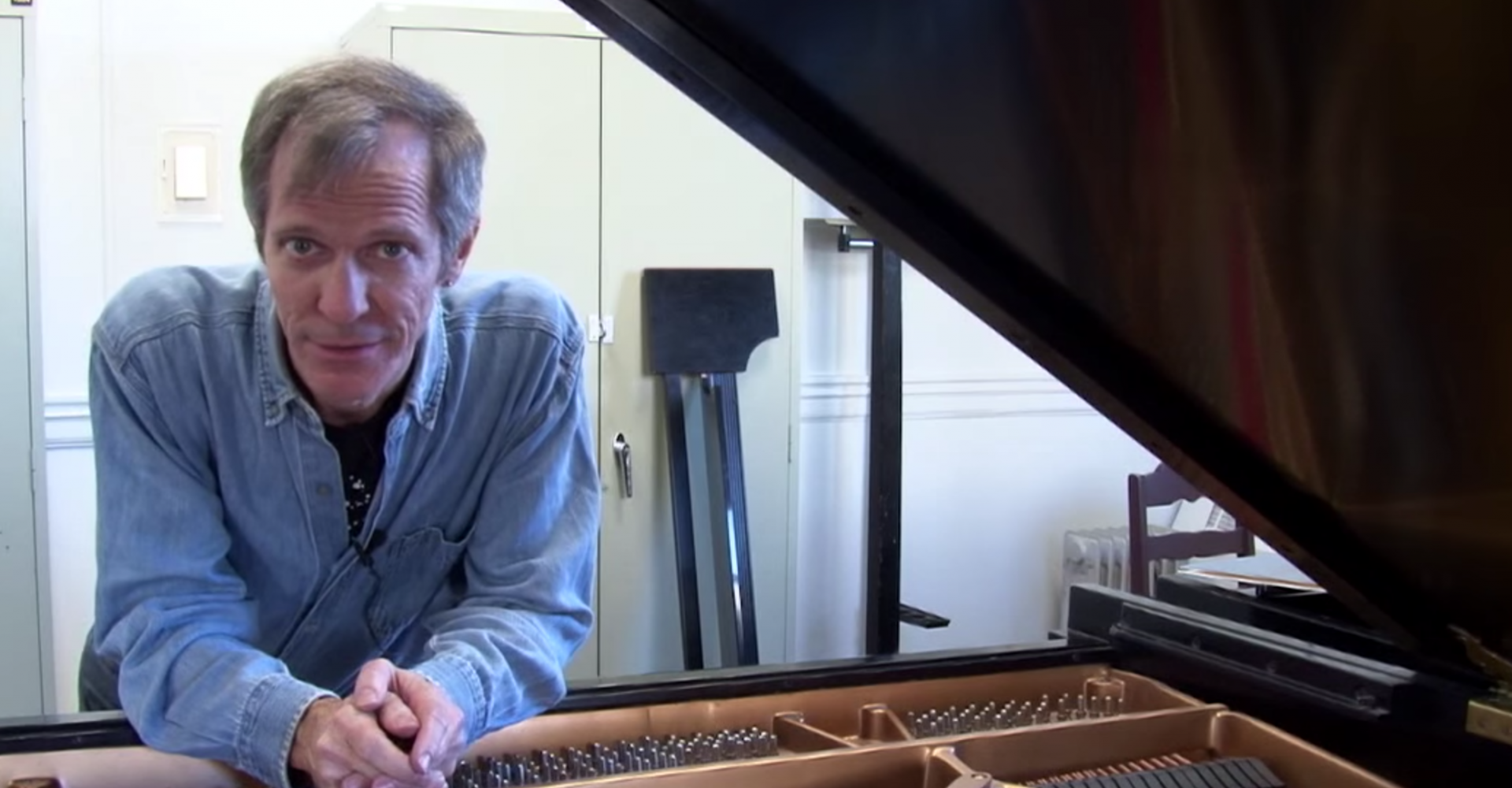 Steve Drury leans on a prepared grand piano looking at the camera