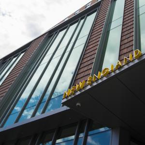 An exterior photo of the Student Life and Performance Center, with the words New England visible and blue sky and clouds visible in the building's reflective windows.