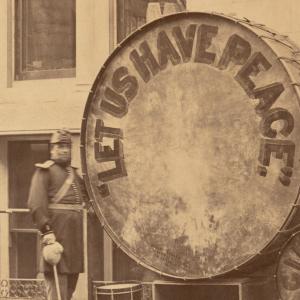 Black and white photo of a man in a band uniform standing near a large drum. The drum says "LET US HAVE PEACE."