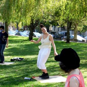 Jenny Herzog smiles while teaching tap in the park. Students are watching her. In the background is bright green grass and trees.